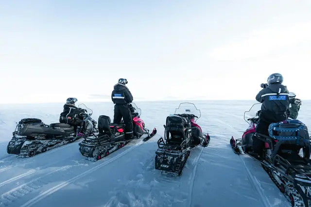 A group of tourists on snowmobiles explore the vast Arctic landscape under clear skies.